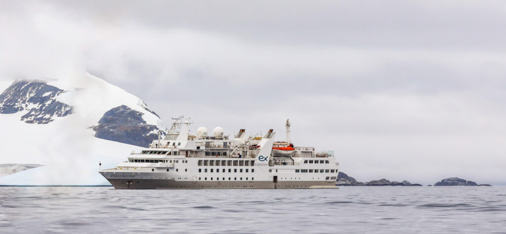 Exploris One expedition ship sailing in Antarctic waters with snowy mountains and cloudy skies in the background